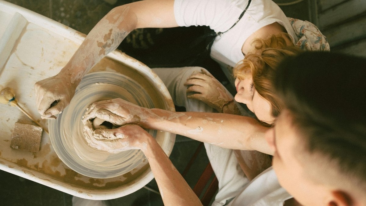 two people with clay-covered hands collaborating on a pottery wheel