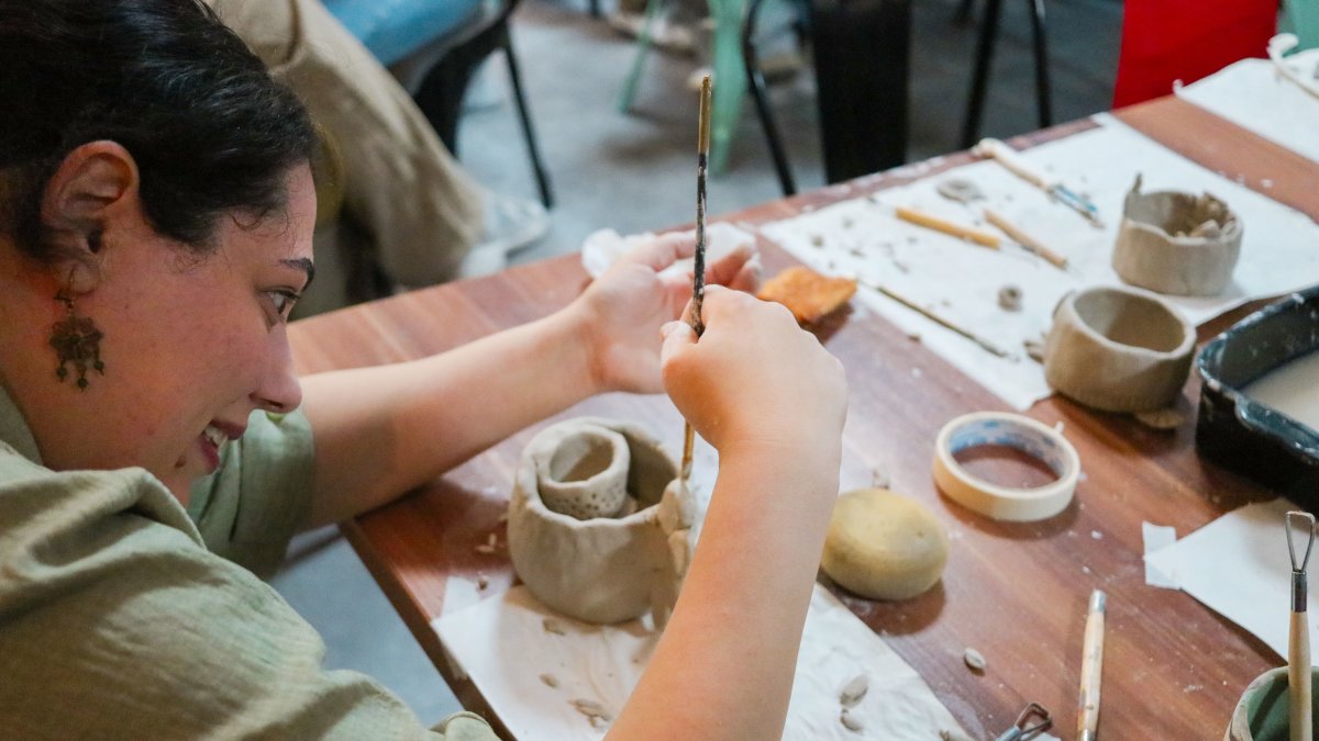 A woman joyfully shapes clay during a pottery workshop, surrounded by crafting tools on a wooden table.