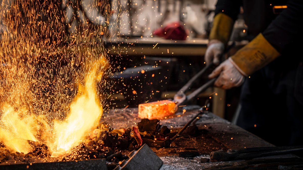 Blacksmith working with hot metal in a fiery forge with sparks flying, showing intense metal shaping process.